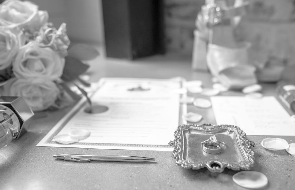 Black and white wedding detail shot featuring rings on silver tray, bouquet, and marriage license on table.