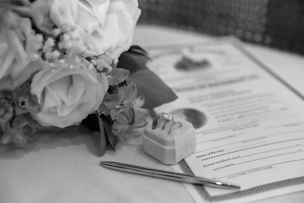 Black and white close-up of wedding rings in box beside bridal bouquet and marriage license document.