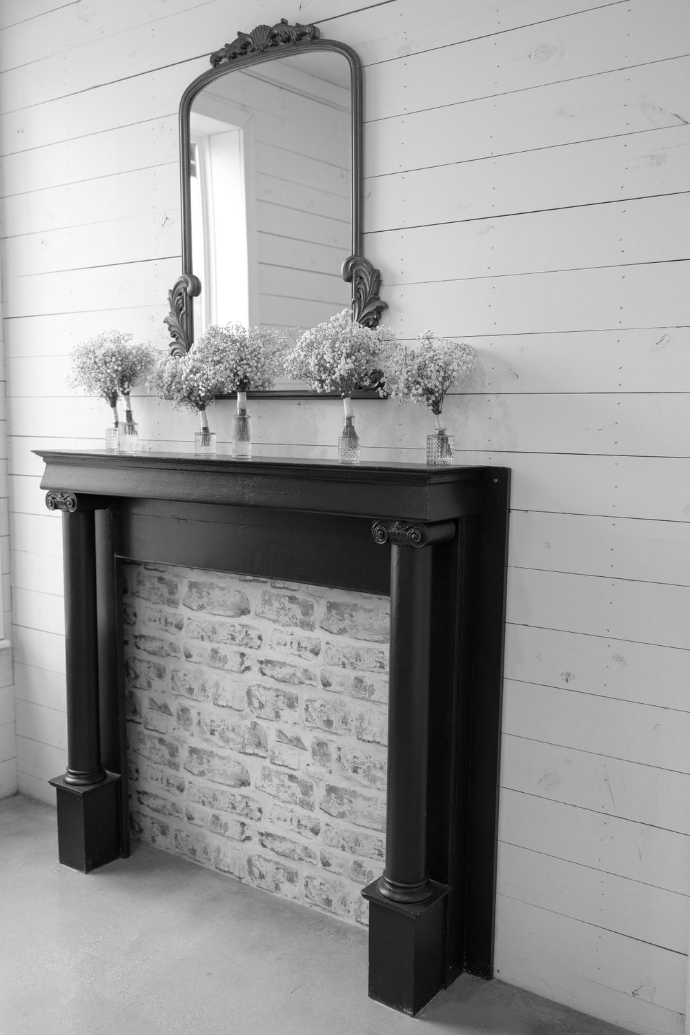 Black and white photo of elegant black fireplace mantel with mirror and baby's breath floral arrangements.