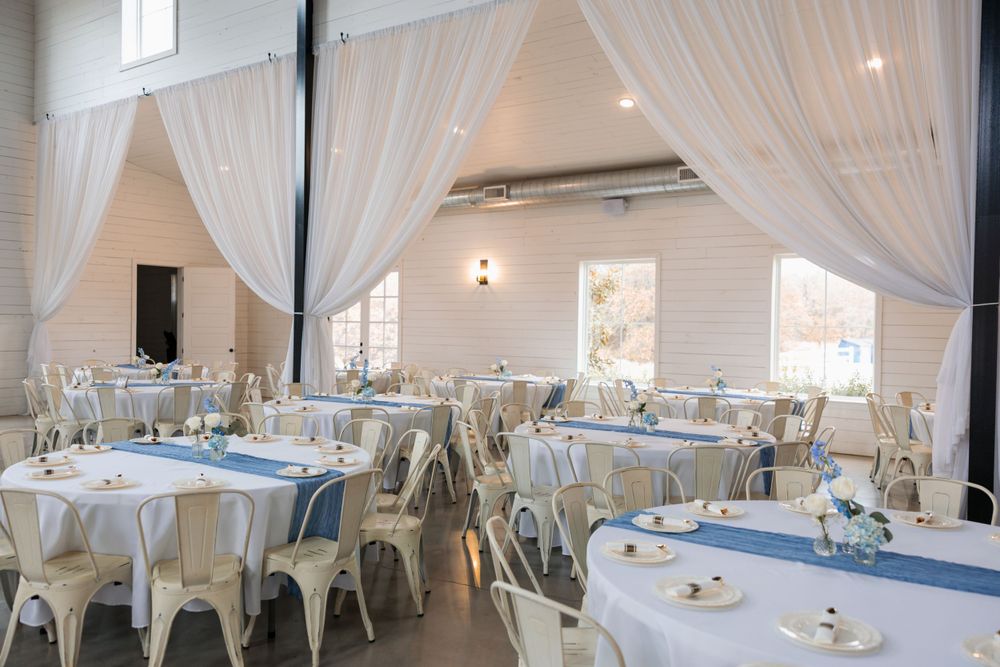 Bright wedding reception venue with white draping, round tables, blue runners, and metal chairs.