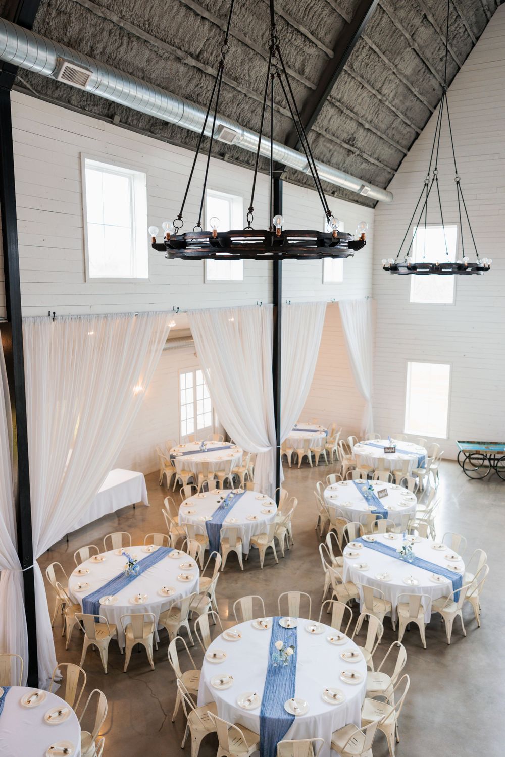 Aerial view of barn wedding reception hall with vaulted ceiling, chandeliers, and blue-accented round tables.