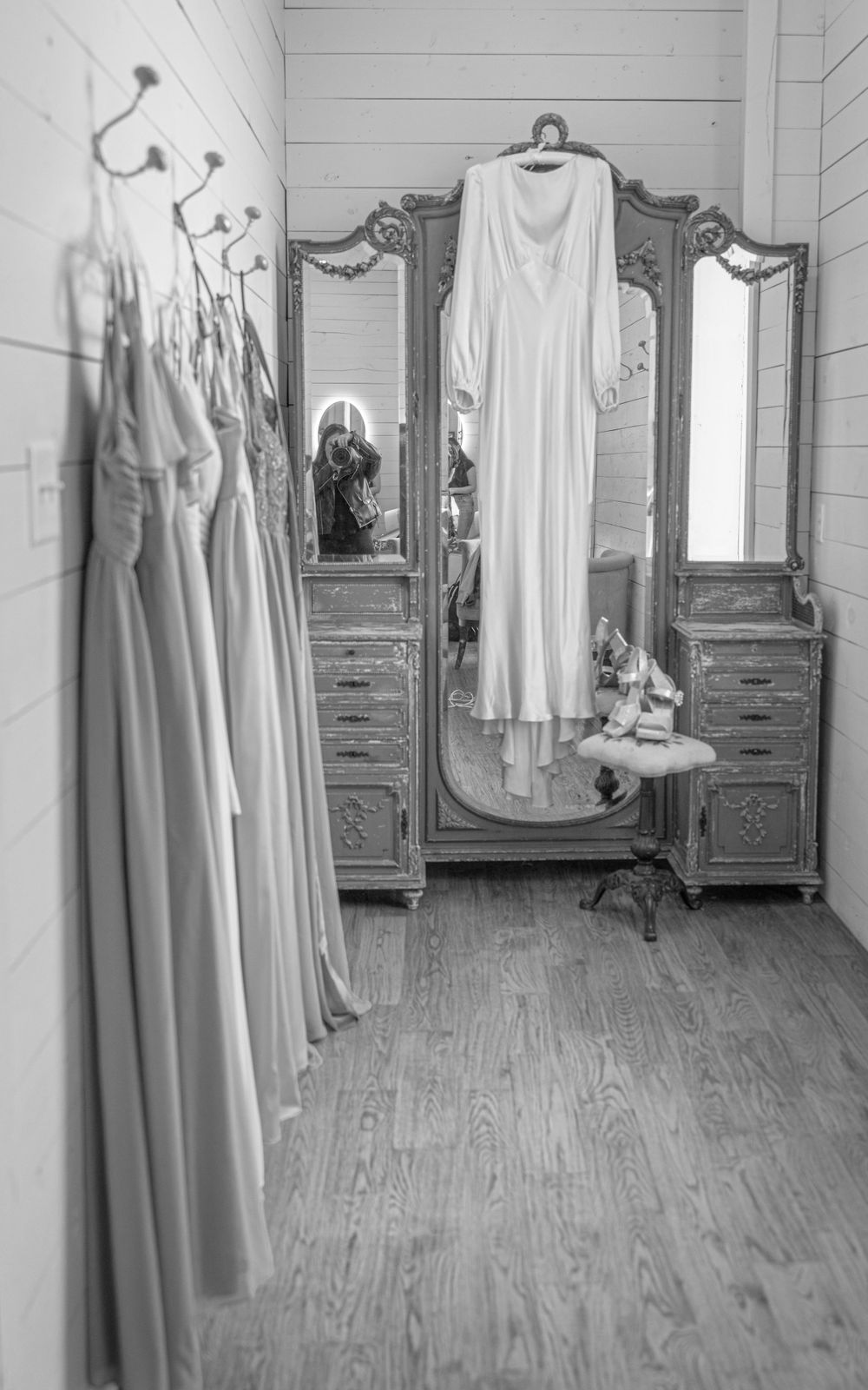 Black and white photo of bridal dressing room with gown on antique mirror and bridesmaid dresses.
