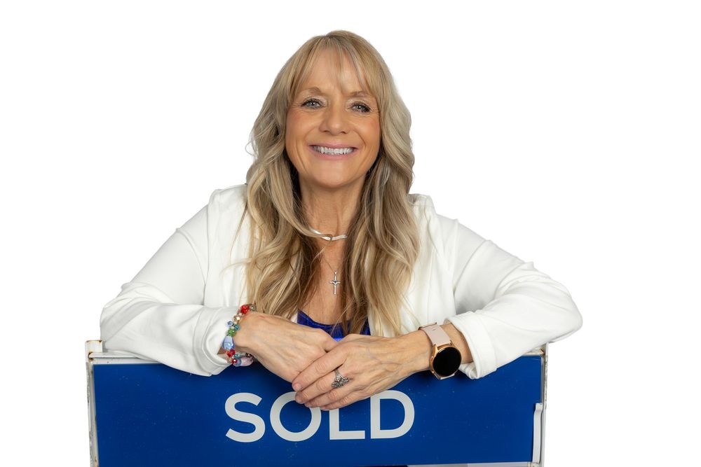 Professional realtor wearing white blazer and blue top poses with SOLD sign for business headshot.