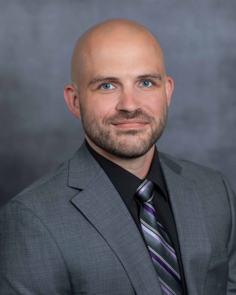 Professional headshot of person in gray suit with purple striped tie against gray background.