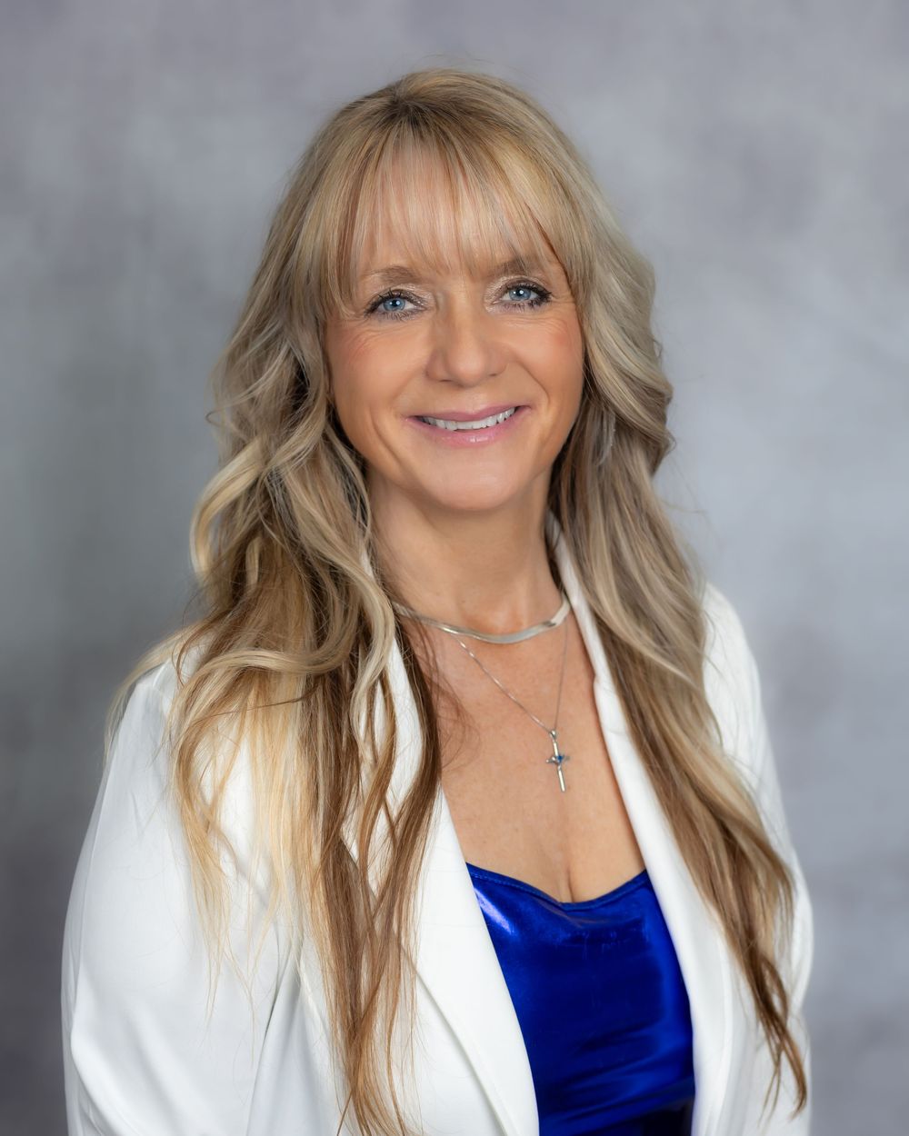 Professional headshot portrait of someone wearing a royal blue top and white blazer against a gray studio backdrop.