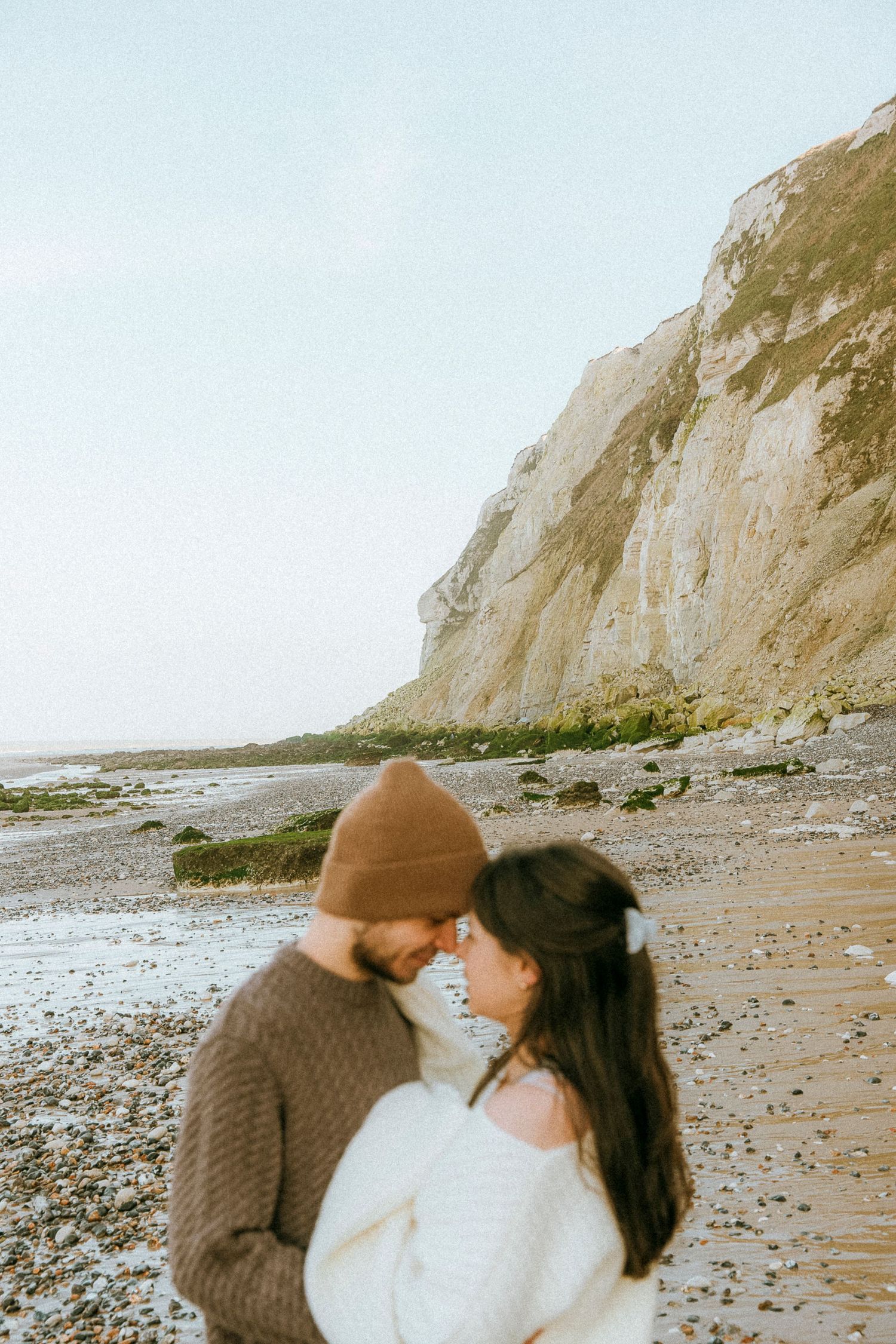 A couple embraces on a rocky beach with towering white cliffs in the background on an overcast day.