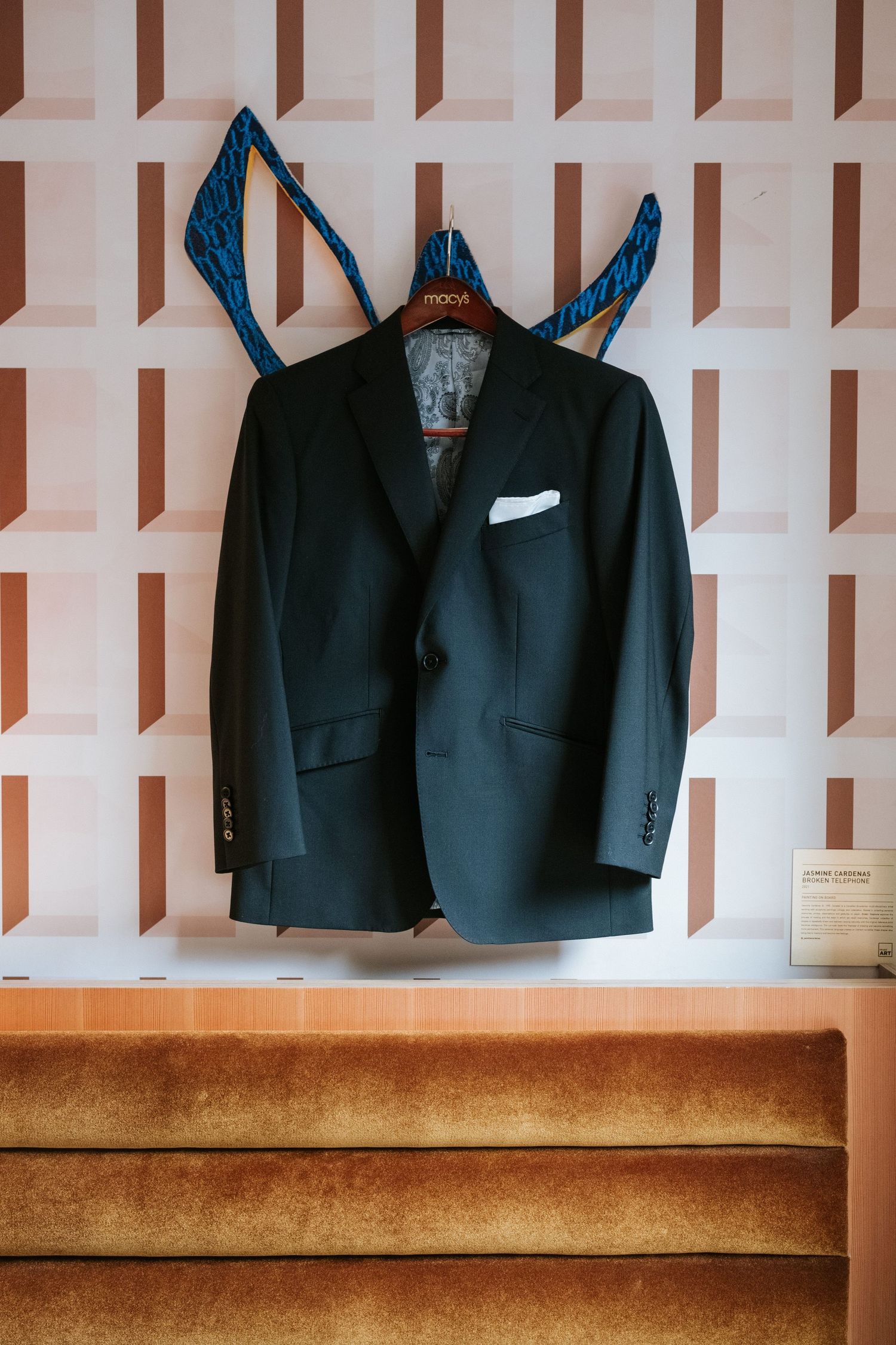 A black formal suit jacket hanging with blue accessories against a geometric pink wall.