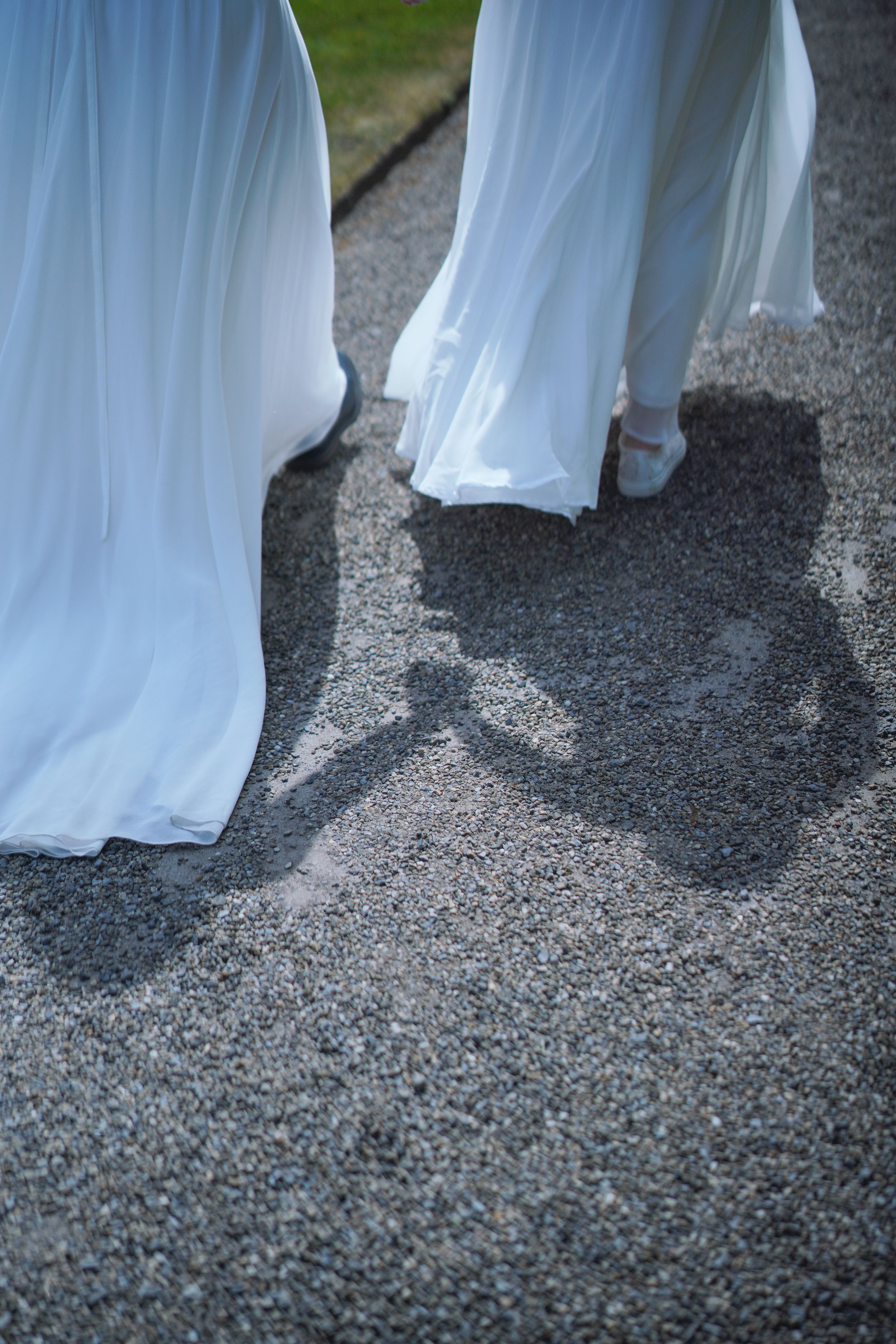 Two women in flowing white bridal gowns walking on gravel path, casting shadows on the ground.
