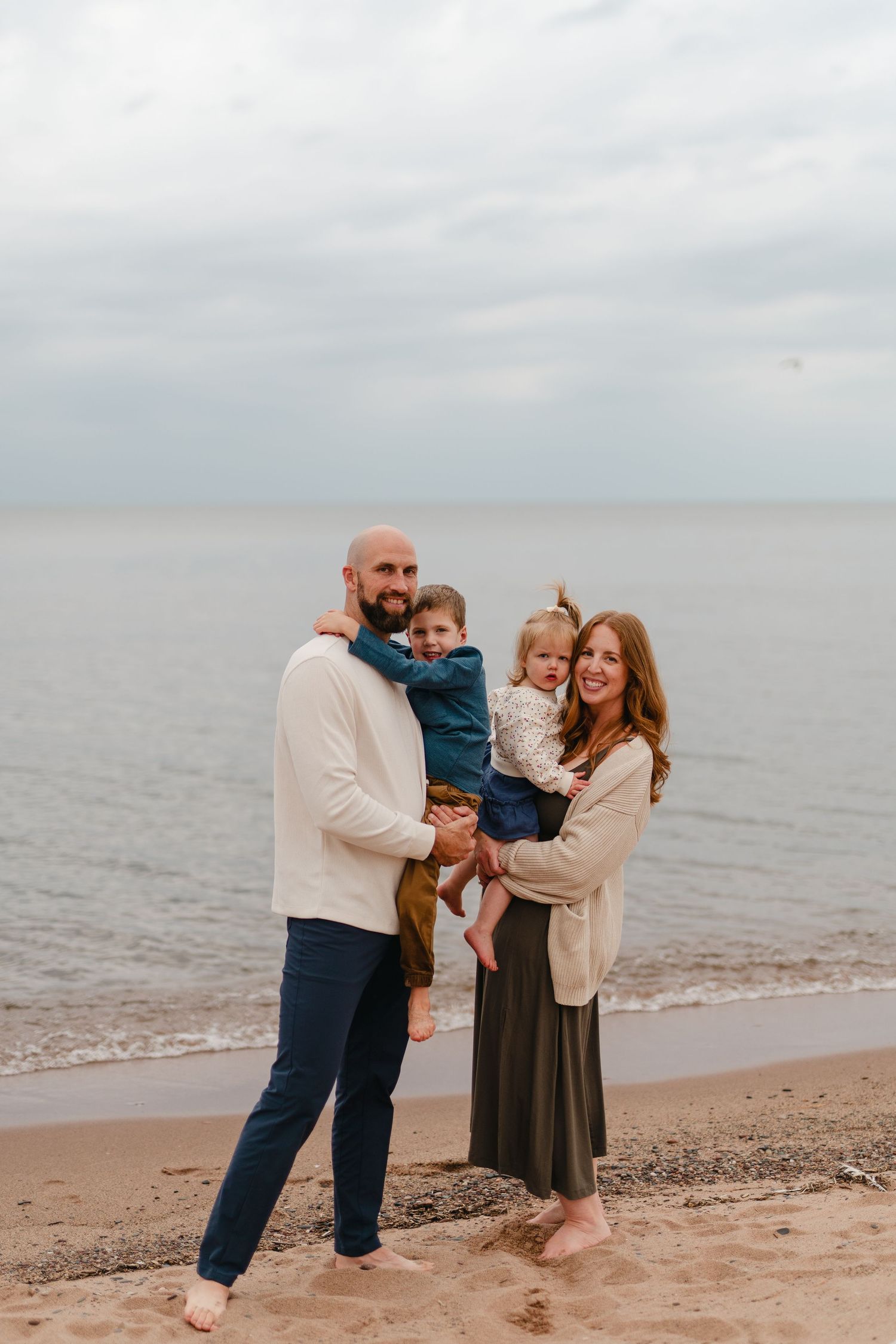 A family wearing cozy sweaters stands together on a cloudy beach against a gray ocean backdrop.