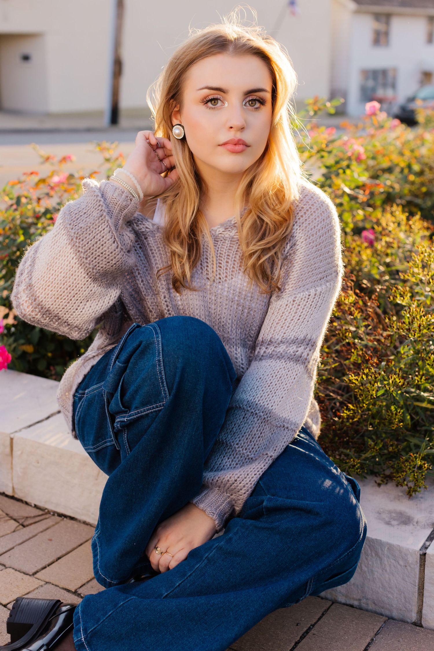 A person in a cozy gray sweater and jeans sits on concrete steps near colorful flowers at sunset.