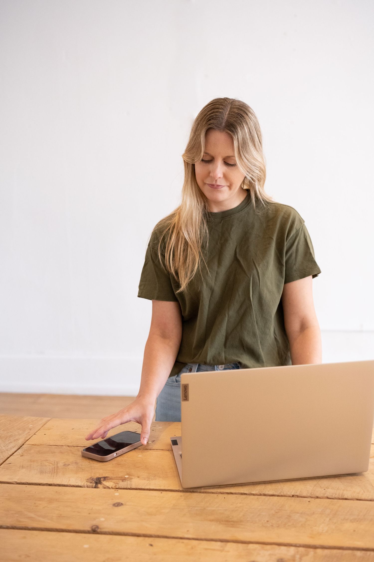 Woman in olive green t-shirt works on laptop at wooden table while reaching for mobile phone.