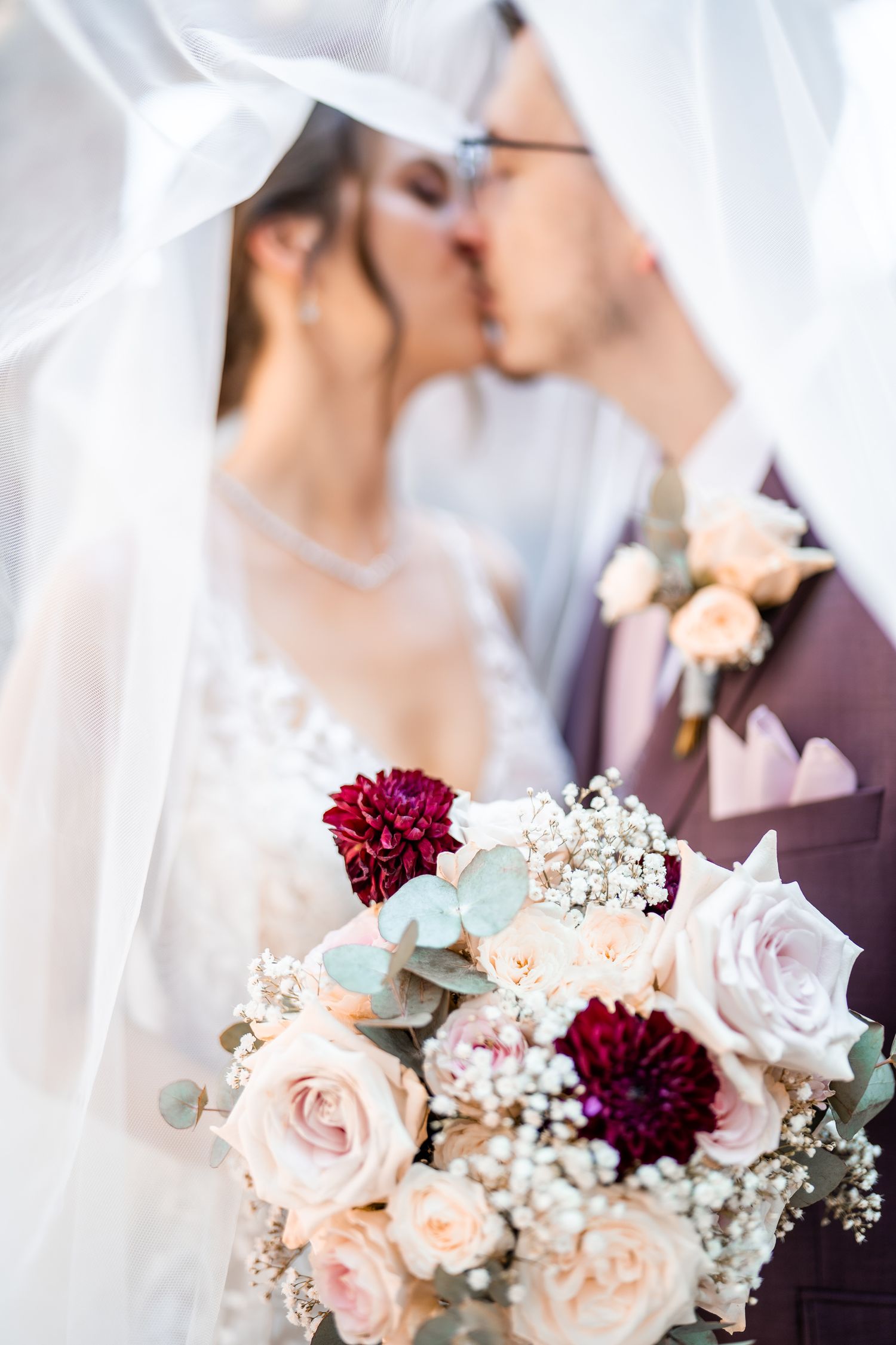 Wedding bouquet with pink and burgundy flowers while couple shares kiss behind bridal veil.