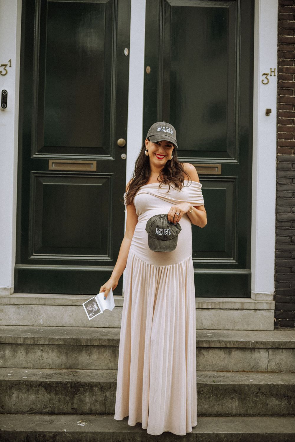 A person in a flowing beige maxi dress and baseball cap stands on stone steps in front of dark green double doors.