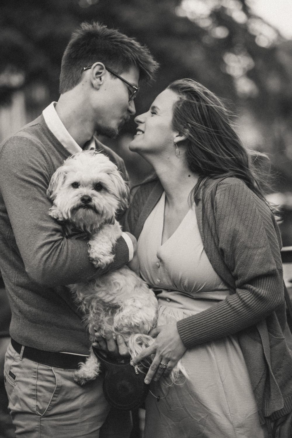 Black and white engagement photo featuring couple with small white dog outdoors.