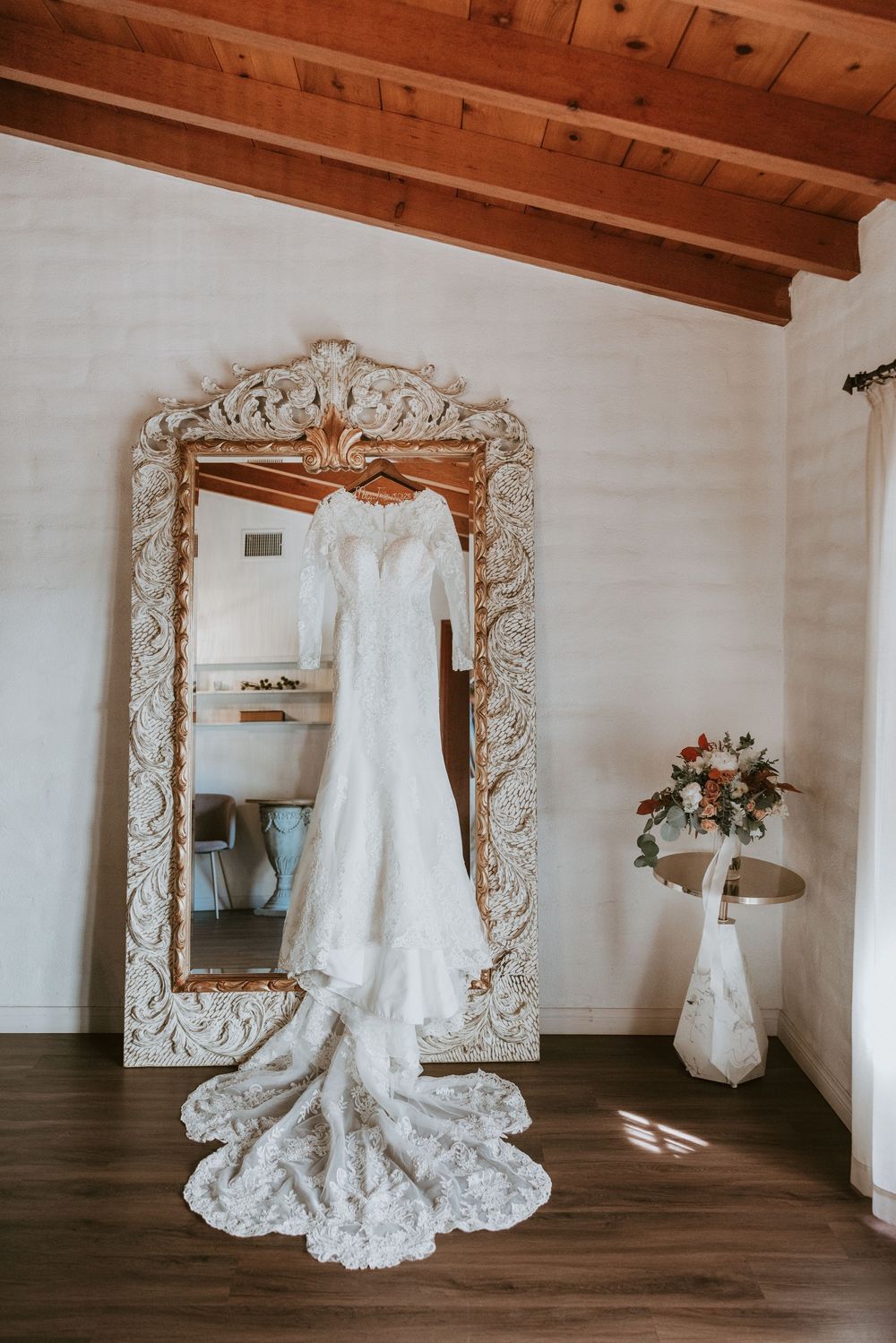 Elegant lace wedding gown with long train hanging on ornate gold mirror in bridal suite.