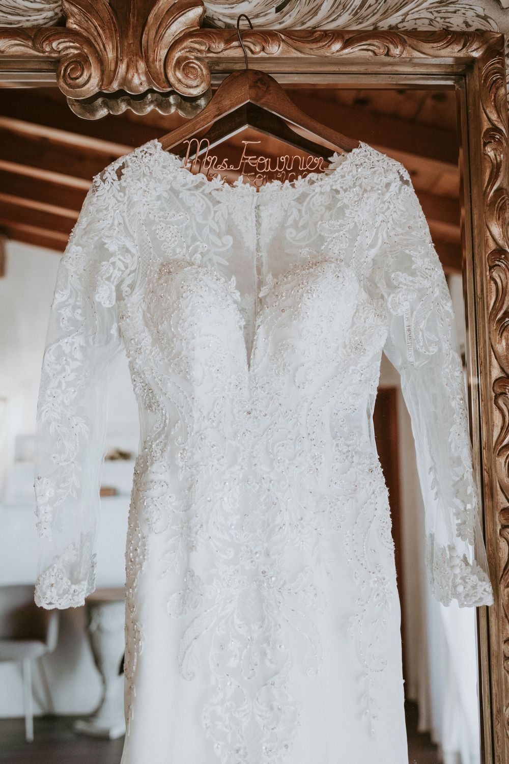 Elegant long-sleeve lace wedding dress hanging on ornate gold mirror frame on custom Mrs. hanger.