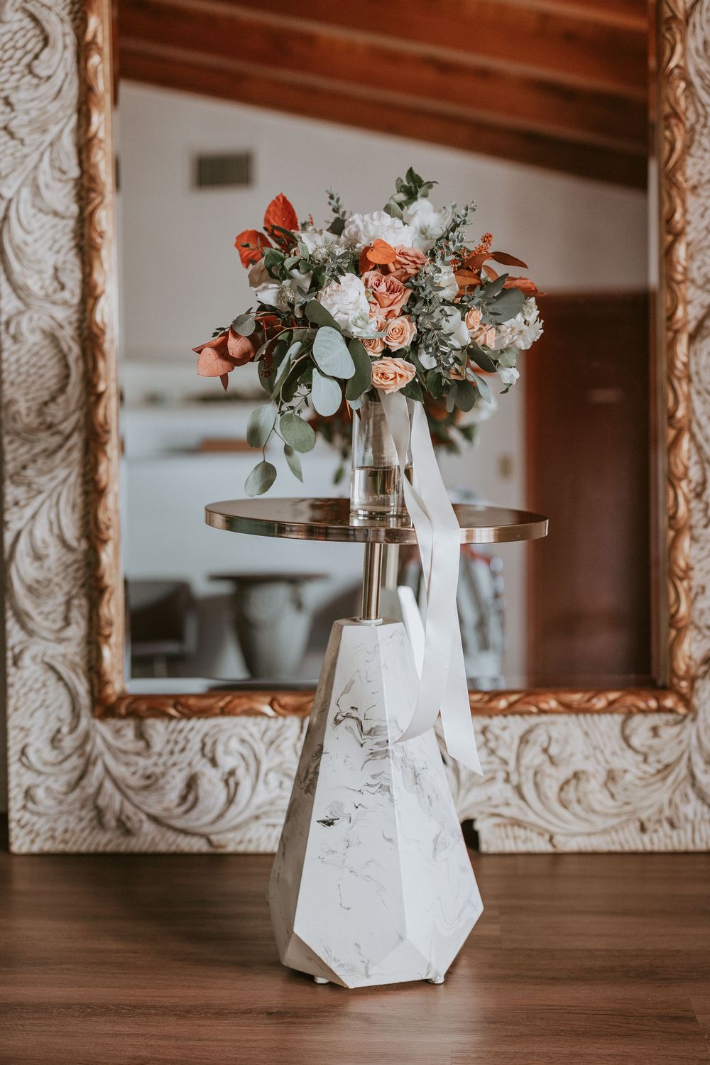 Romantic bridal bouquet with peach and white flowers displayed on marble pedestal by ornate mirror.