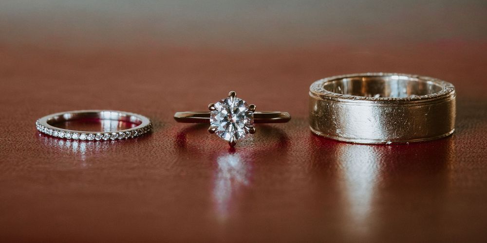 Three wedding rings displayed on a wooden surface, including a diamond engagement ring, eternity band, and gold mens band.