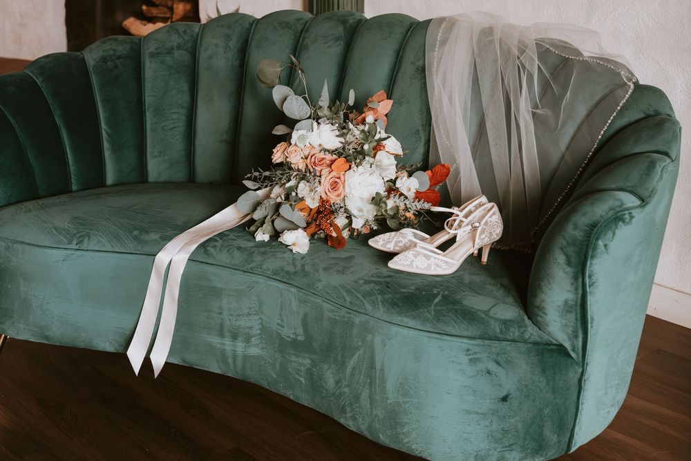 Bridal bouquet of orange and white flowers with veil and heels on teal velvet sofa.