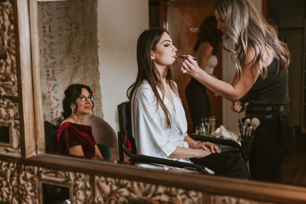 Bride gets makeup applied by artist while bridesmaid watches in ornate mirror setting.