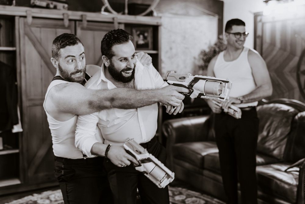 Black and white photo of groomsmen playfully posing with bow ties before wedding ceremony.