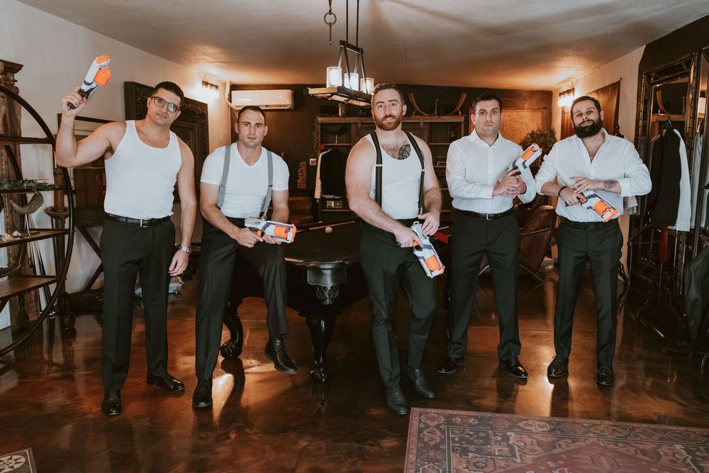 Five groomsmen in white shirts and black suspenders pose playfully indoors holding colorful drinks before a wedding.