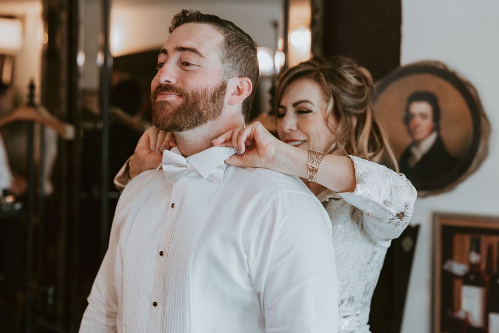 Bride helping groom adjust his bow tie while smiling in a warmly decorated getting-ready room.