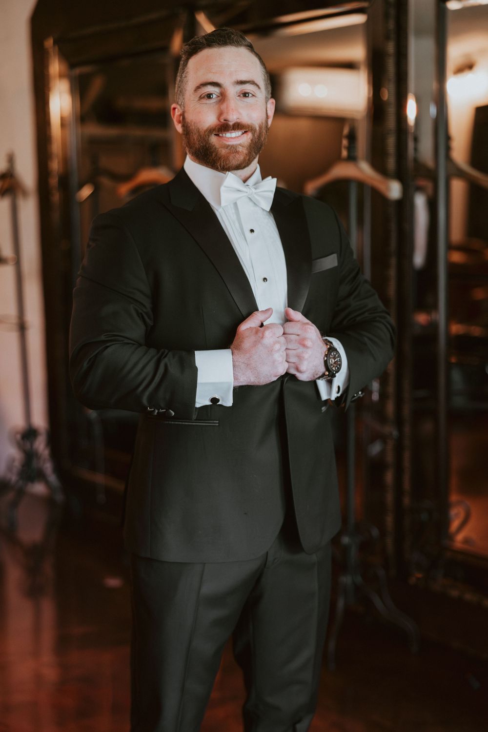 Handsome groom in black tuxedo with white bow tie smiling during wedding day preparations.