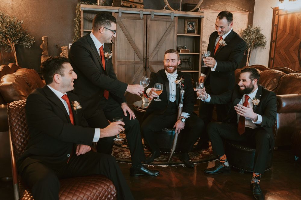 Groom and groomsmen in black tuxedos toast with drinks in a rustic lounge.