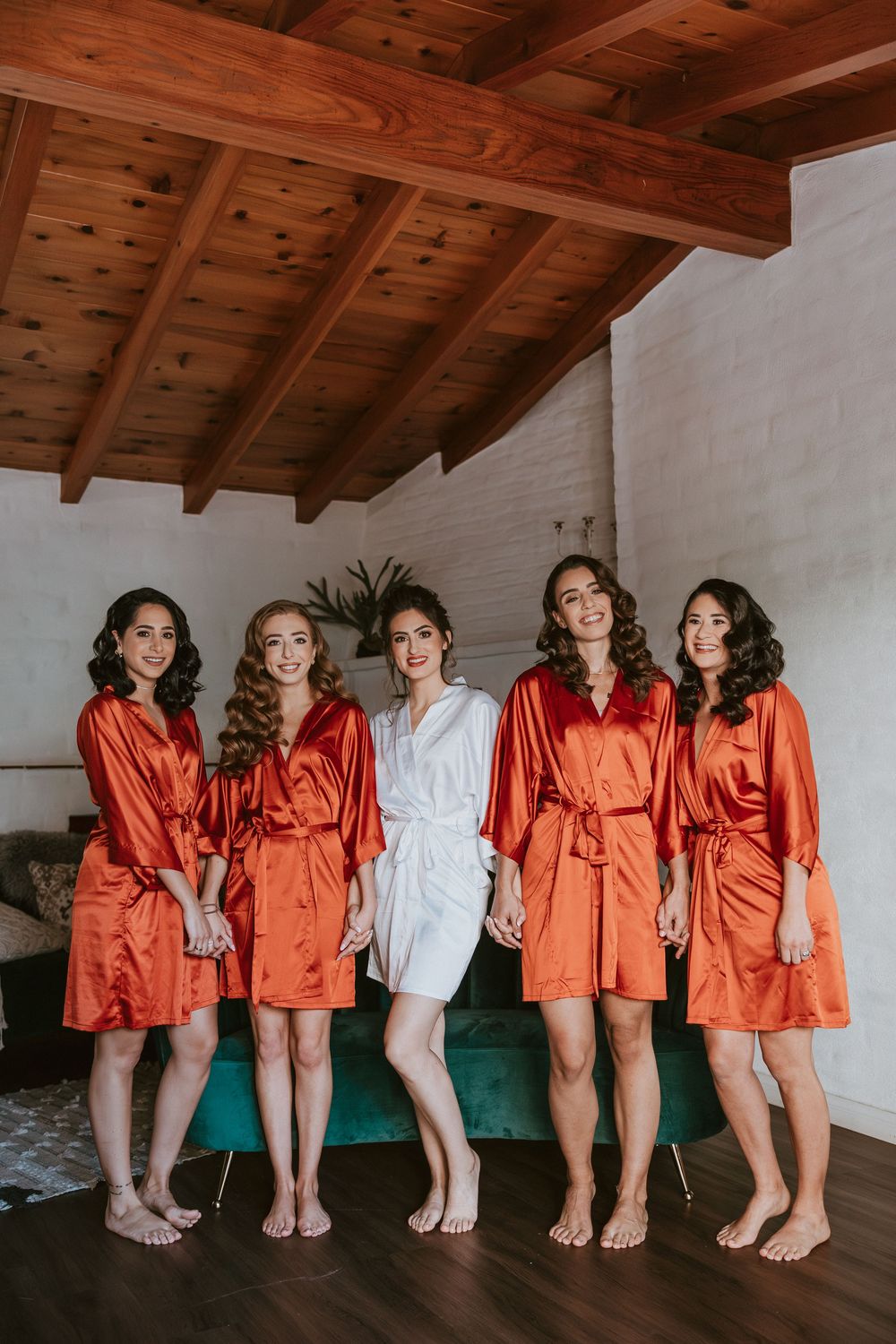 Bride in white robe poses with four bridesmaids in matching orange satin robes indoors.