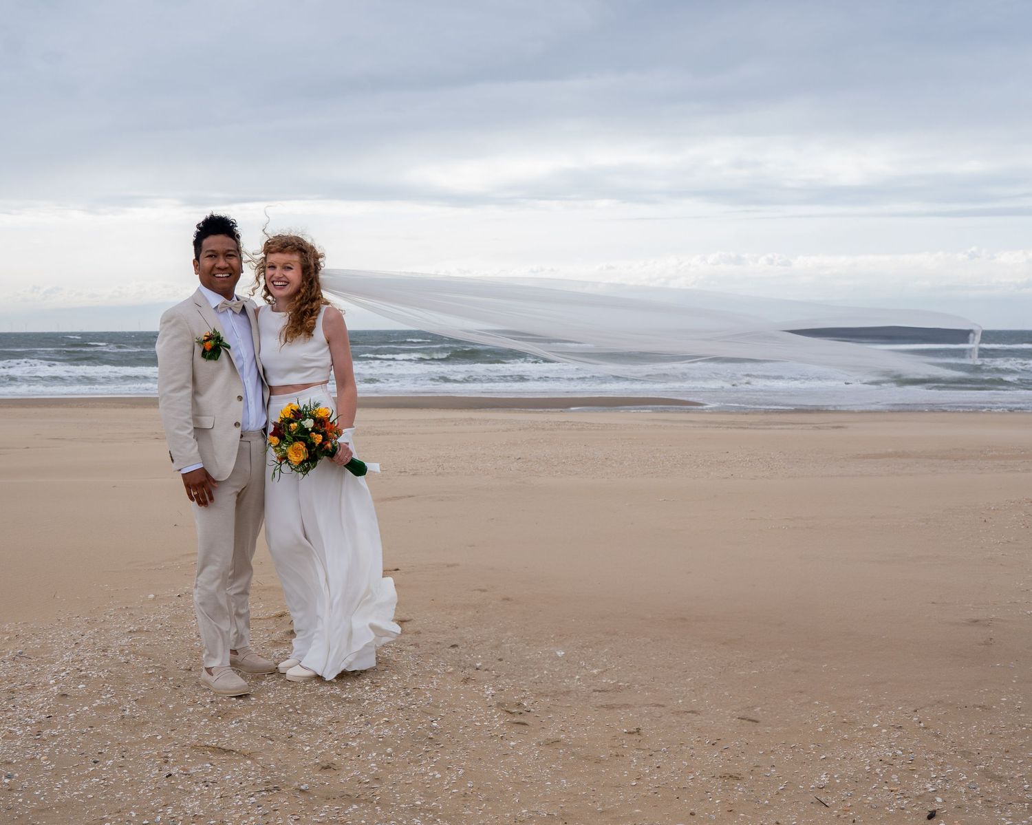 Een stralend bruidspaar poseert samen op een zandstrand met de oceaan op de achtergrond. Trouwfotograaf Heerde