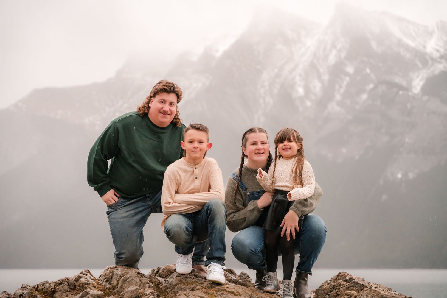Family of four posing on rocks with misty mountain and lake backdrop at Lake Minnewanka, Banff.