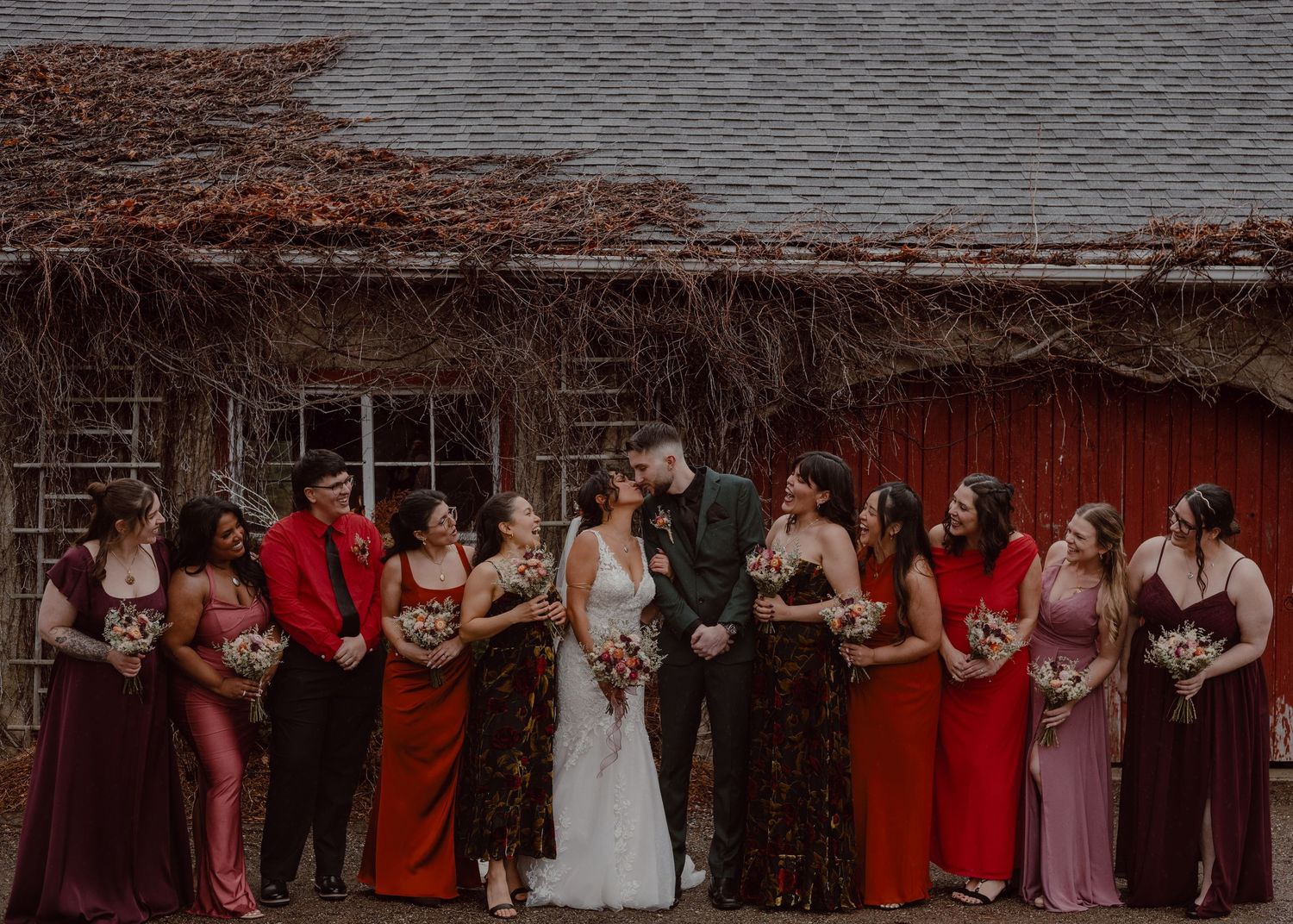 Bride and groom share a kiss in front of a carriage house while the bridesmaids all sneak a peak.