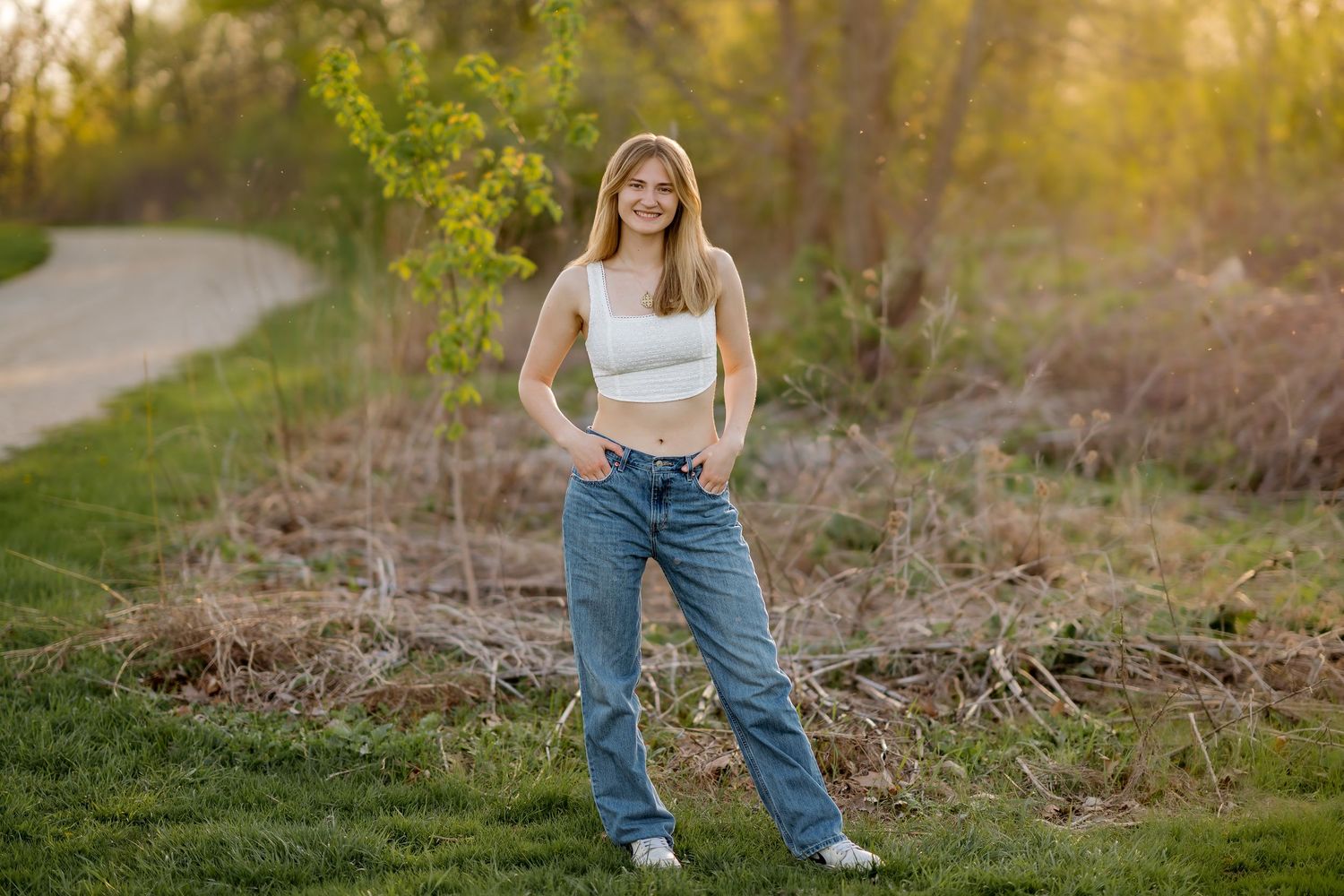 Young blonde woman smiling in white crop top and jeans, posing outdoors in a lush green park at golden hour.