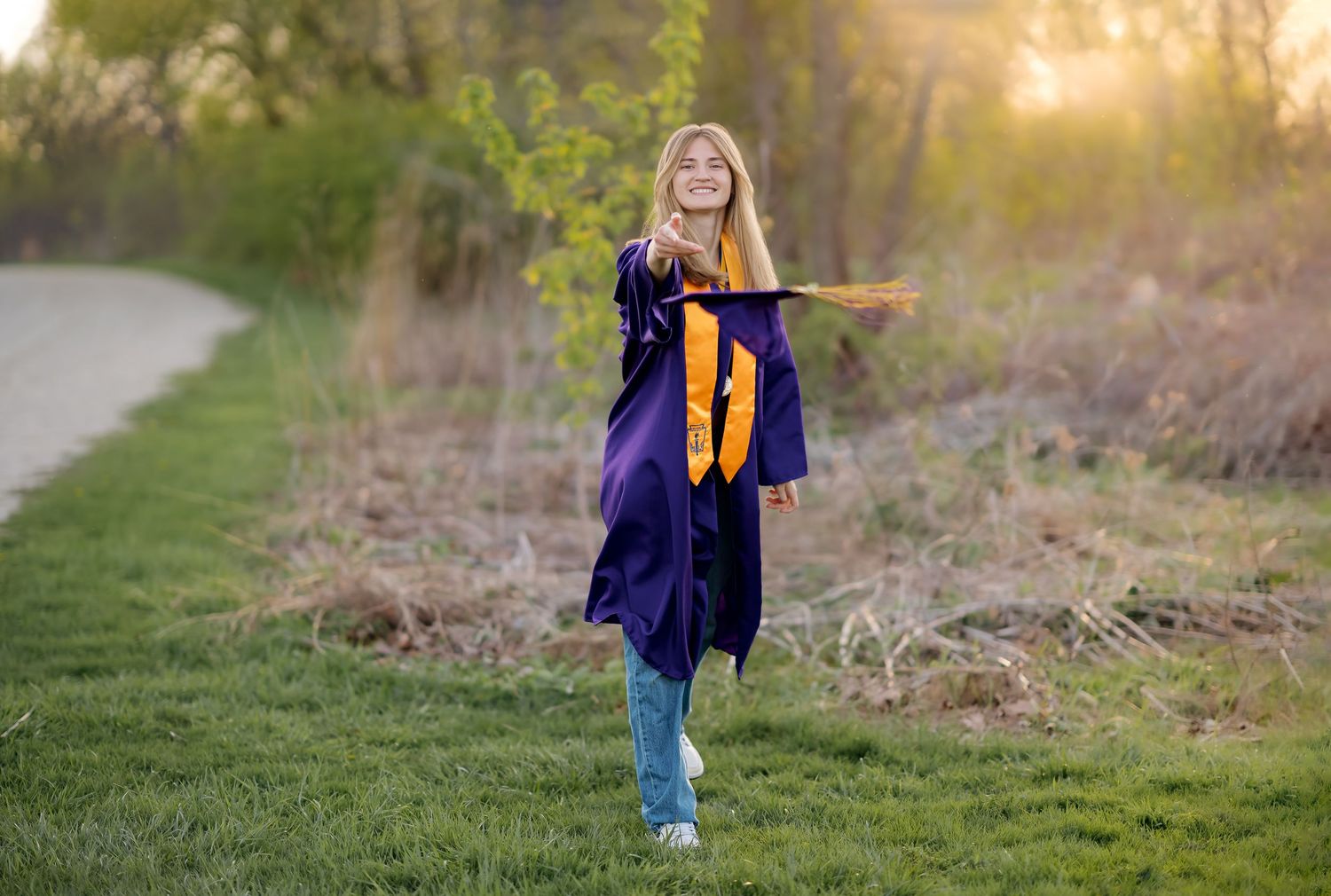 Young woman in purple and gold graduation gown walks outdoors on green grass at sunset, arms outstretched.