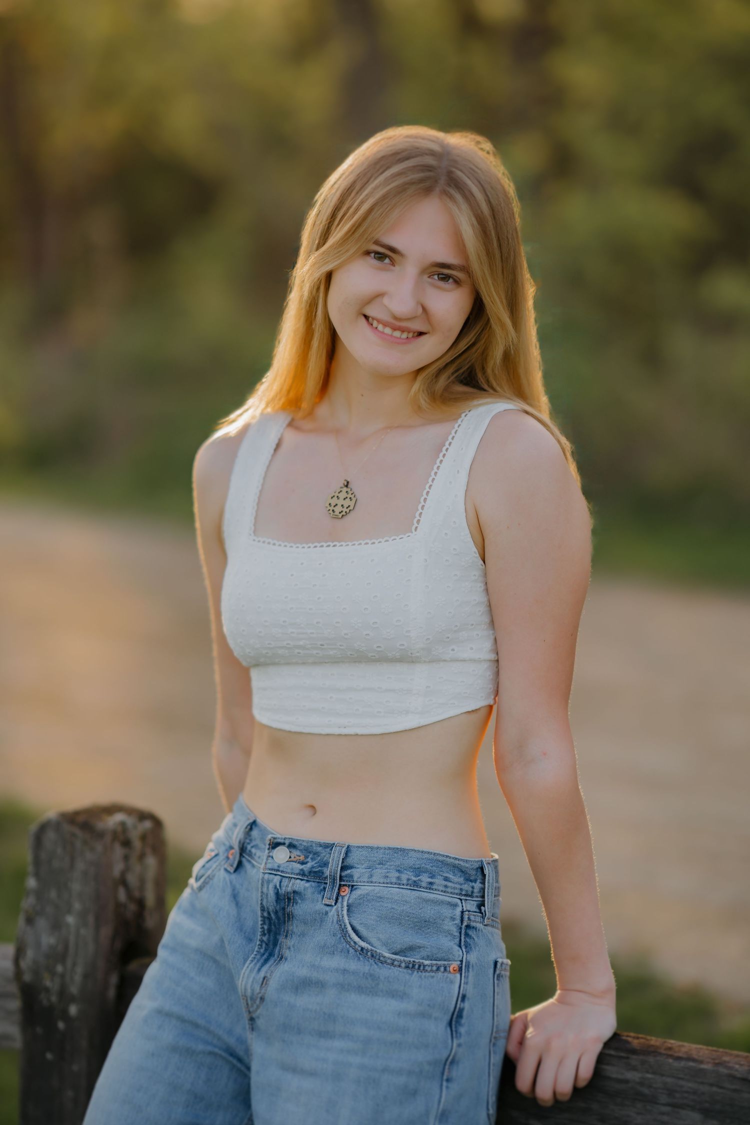 Young woman with blonde hair smiling outdoors, wearing white crop top and jeans, leaning on wooden fence.
