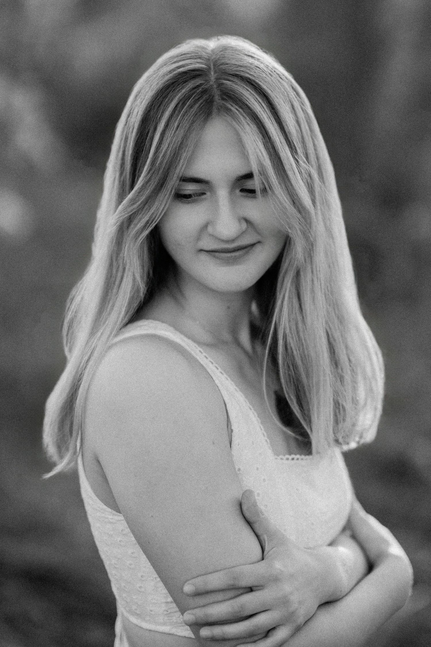 Black and white portrait of a young blonde woman in a white dress, smiling softly with arms crossed outdoors.