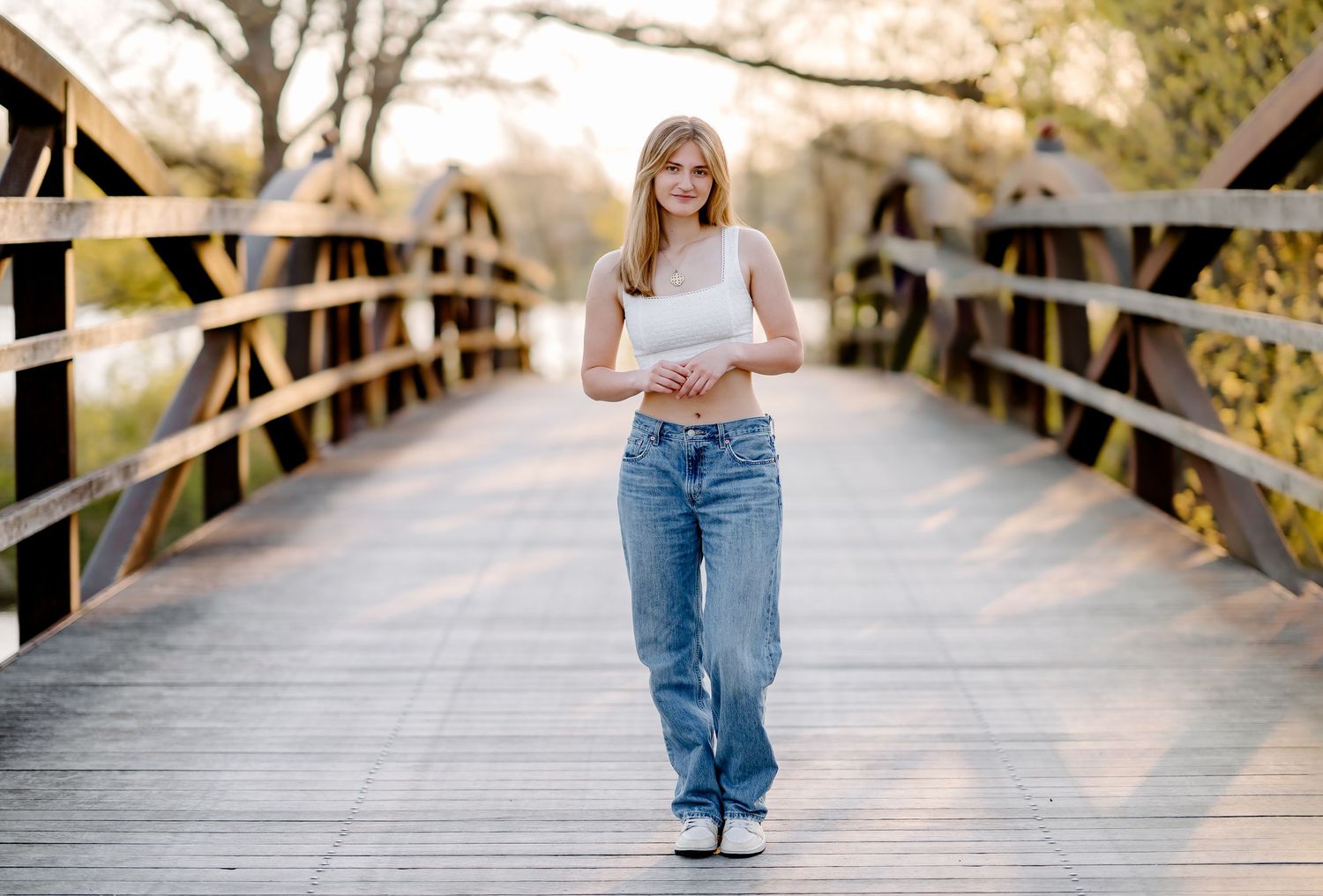 Young blonde woman in white crop top and jeans walking on a wooden bridge surrounded by trees.
