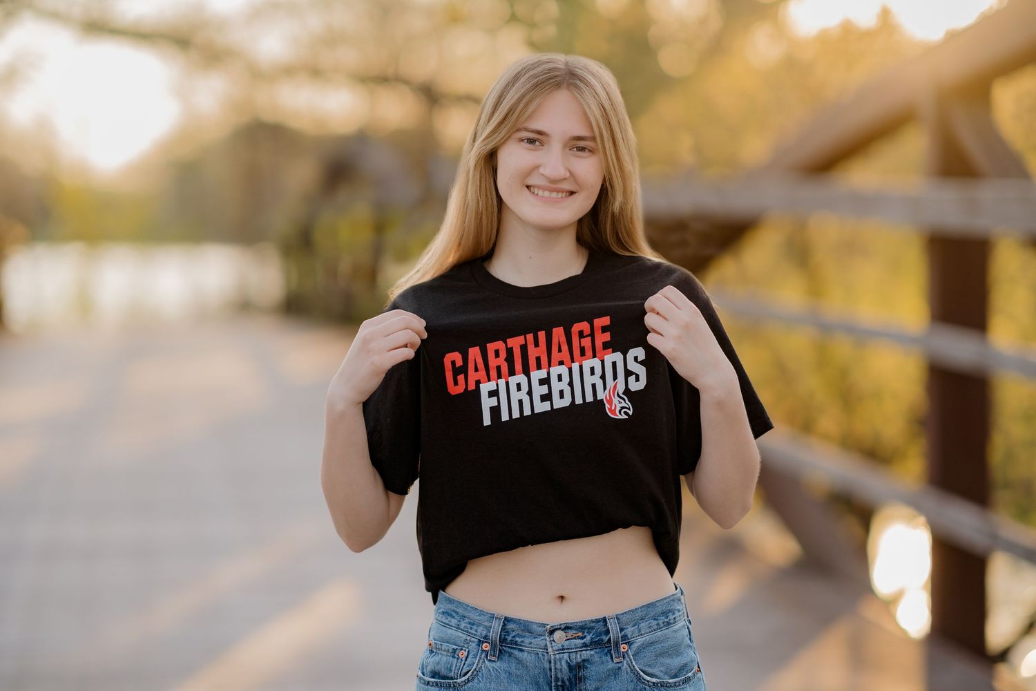 Young woman proudly displaying a black Carthage Firebirds t-shirt outdoors on a wooden bridge.