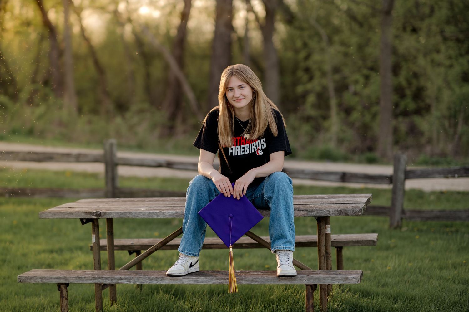 Smiling graduate in black Firebirds shirt sits on picnic table holding purple graduation cap outdoors.