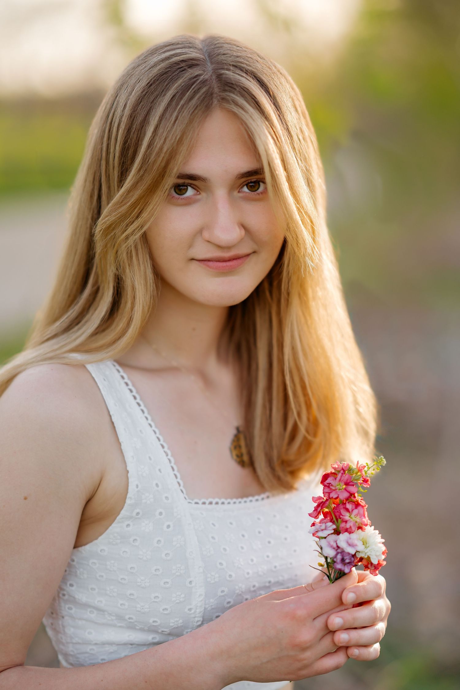 Young blonde woman in white dress holding pink and white wildflowers outdoors in golden sunlight.