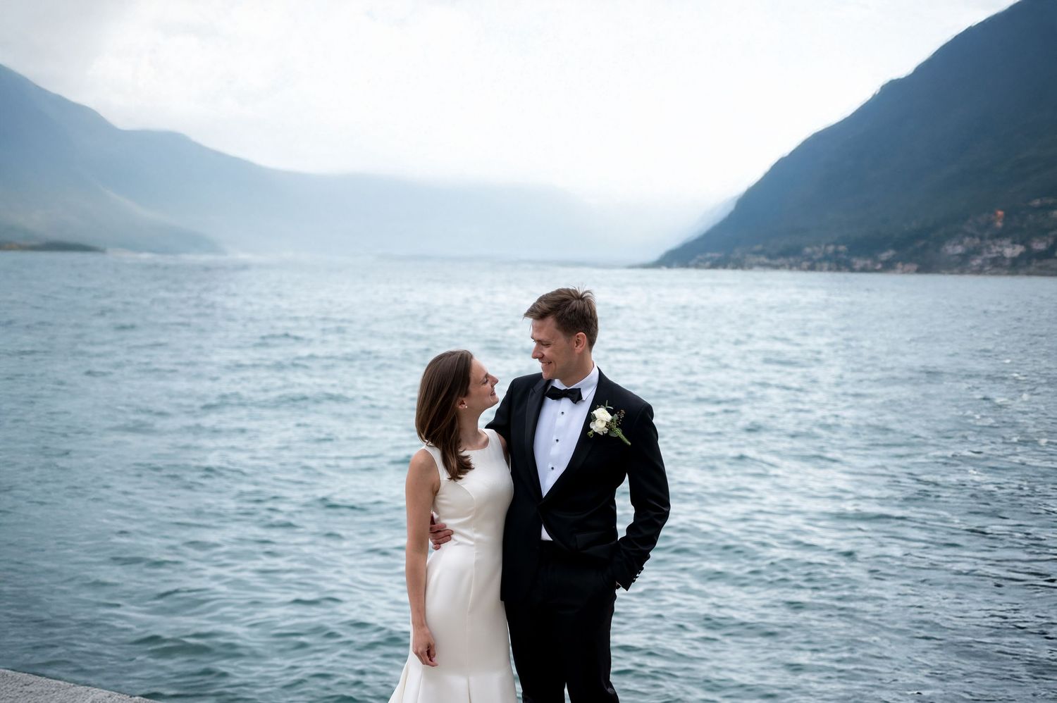 Bride and groom share a romantic moment by a serene lake with mountain backdrop on their wedding day.