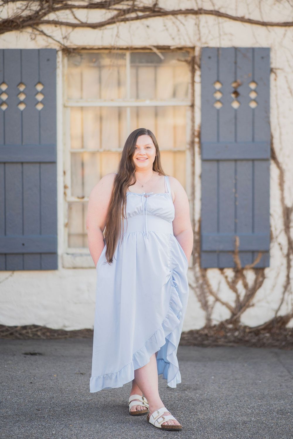 Young woman in light blue ruffled maxi dress standing outdoors near rustic white building with gray shutters.