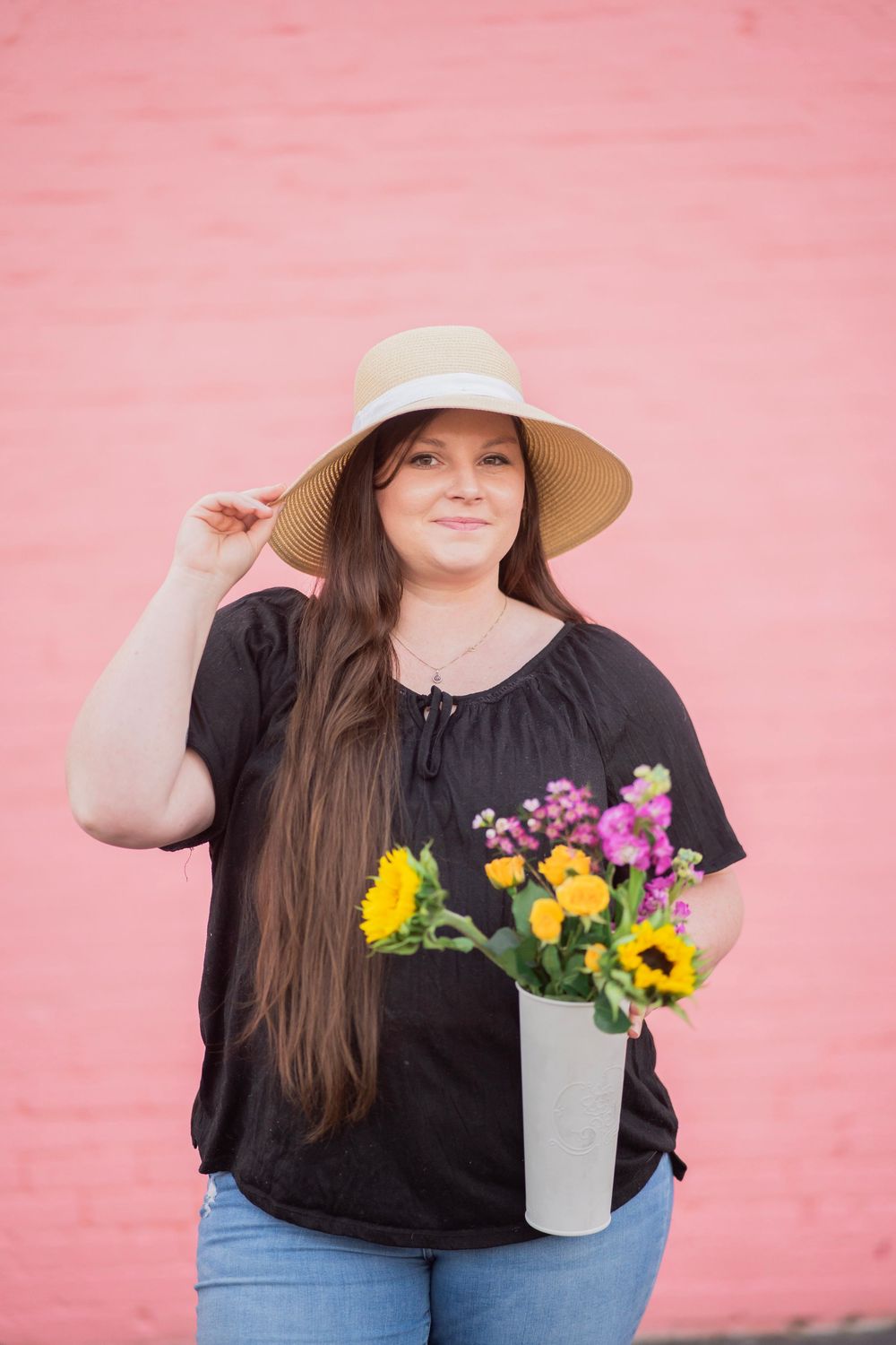 Smiling young woman in black top and sun hat holds a white vase with sunflowers against a pink wall.