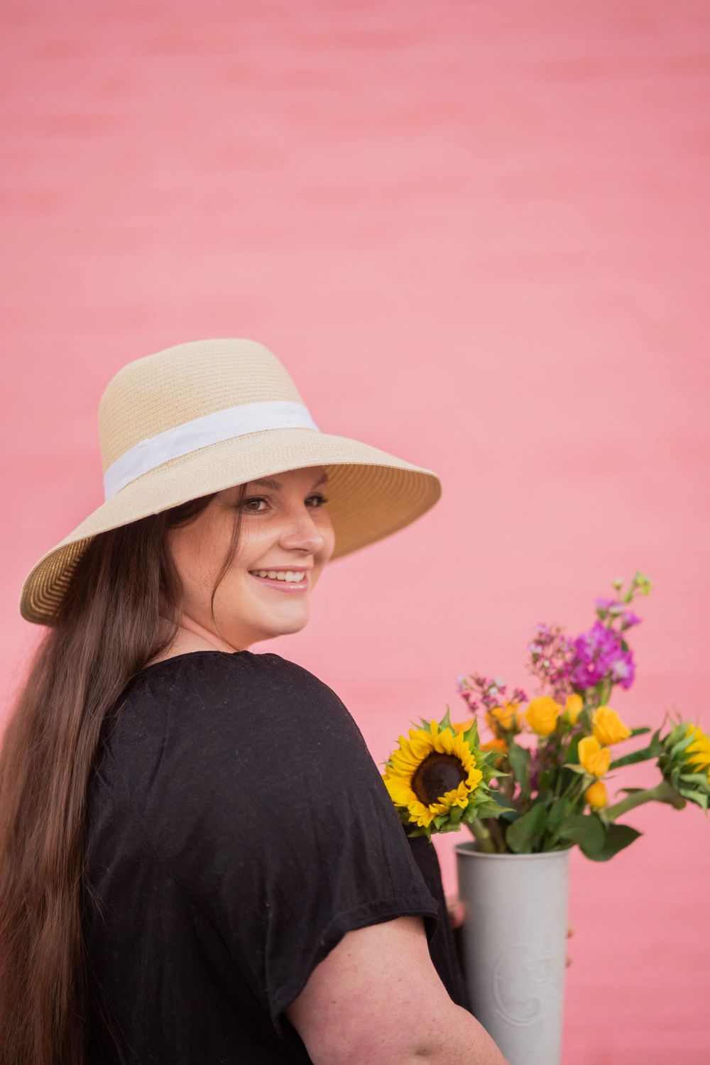 Young woman in straw hat smiles over her shoulder while holding a vase of colorful flowers by a pink wall.