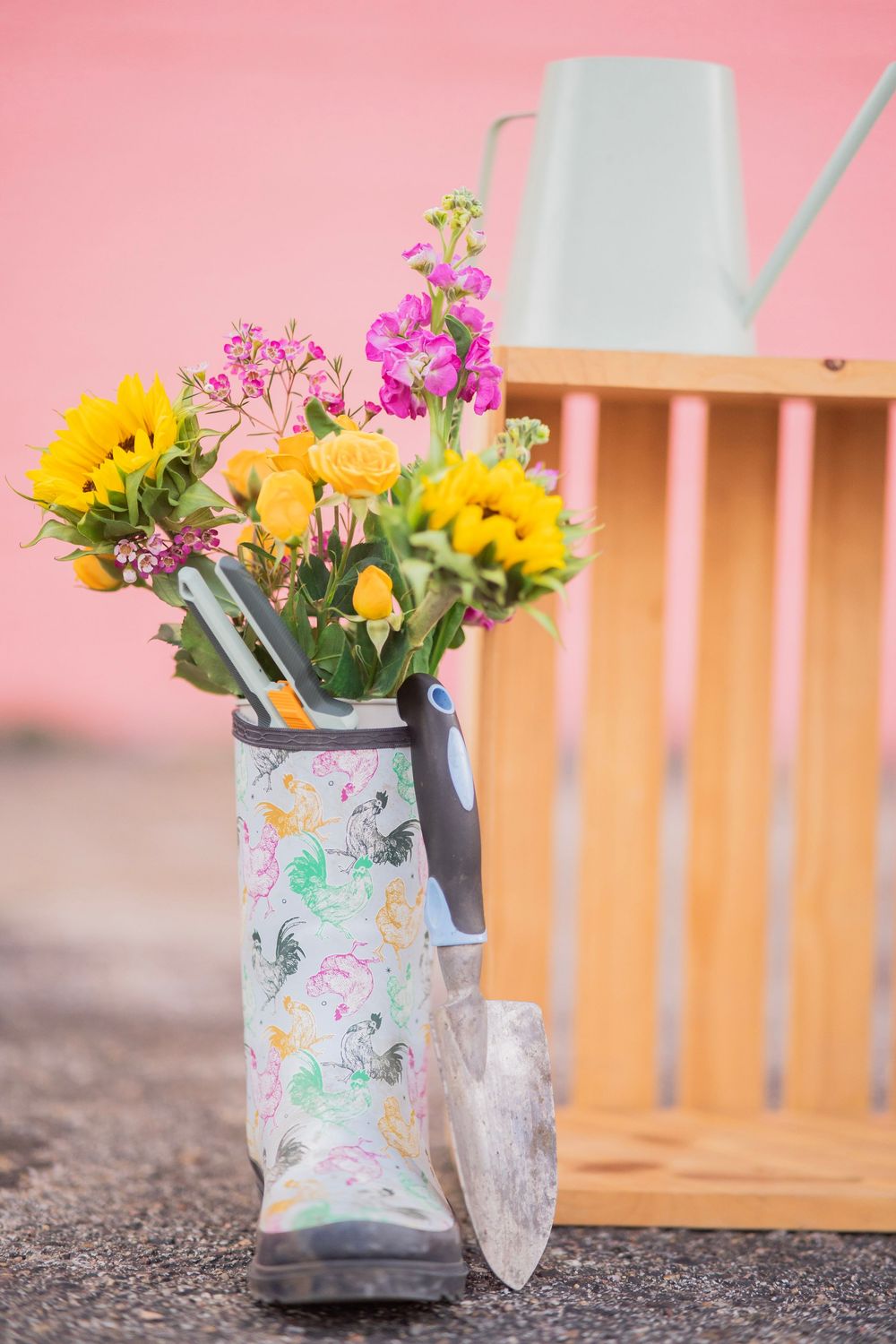 Colorful flowers and garden tools arranged in a patterned rain boot beside a wooden crate on pink background.