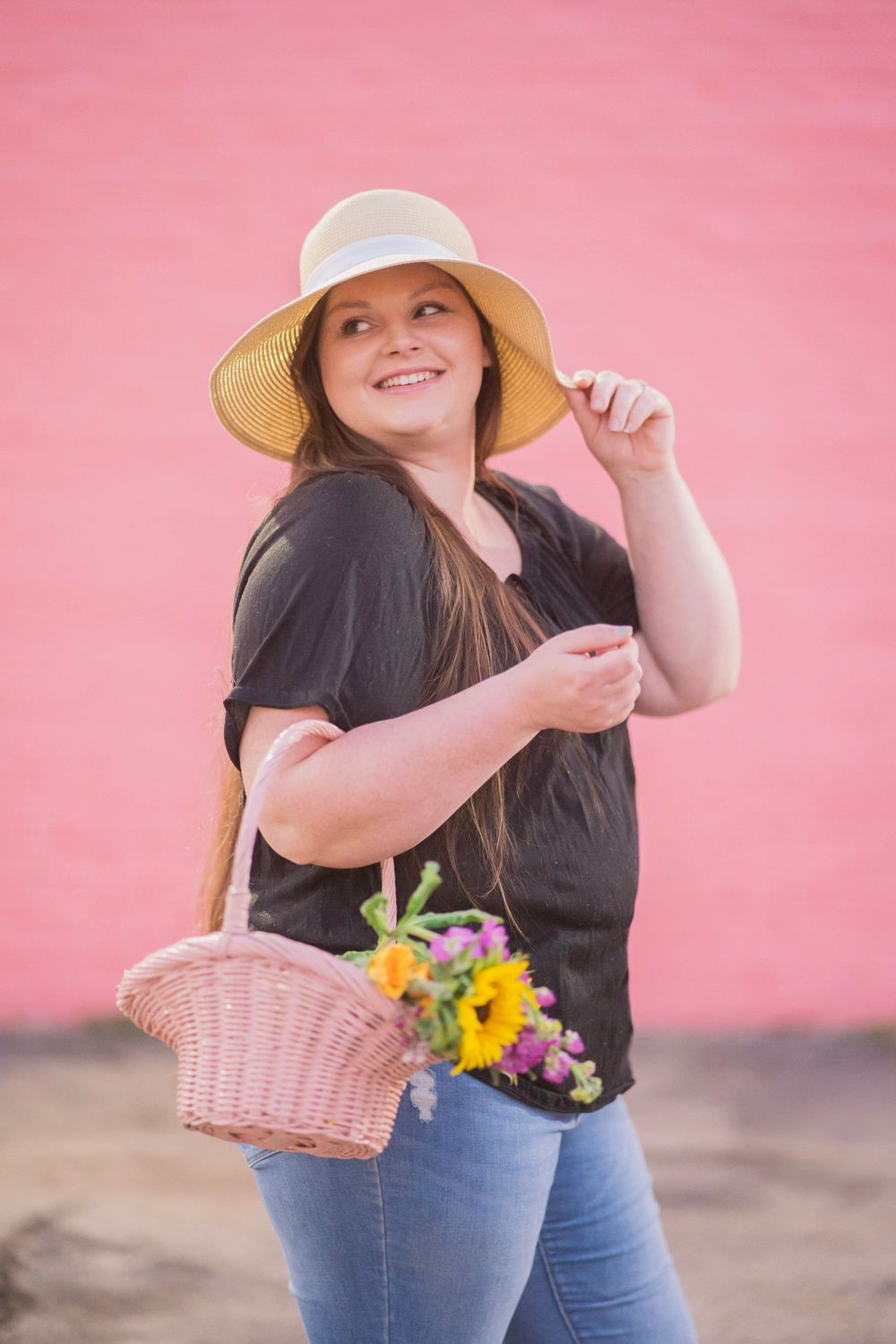 Cheerful young woman in sun hat and jeans carries a pink wicker basket with sunflowers against a pink wall.