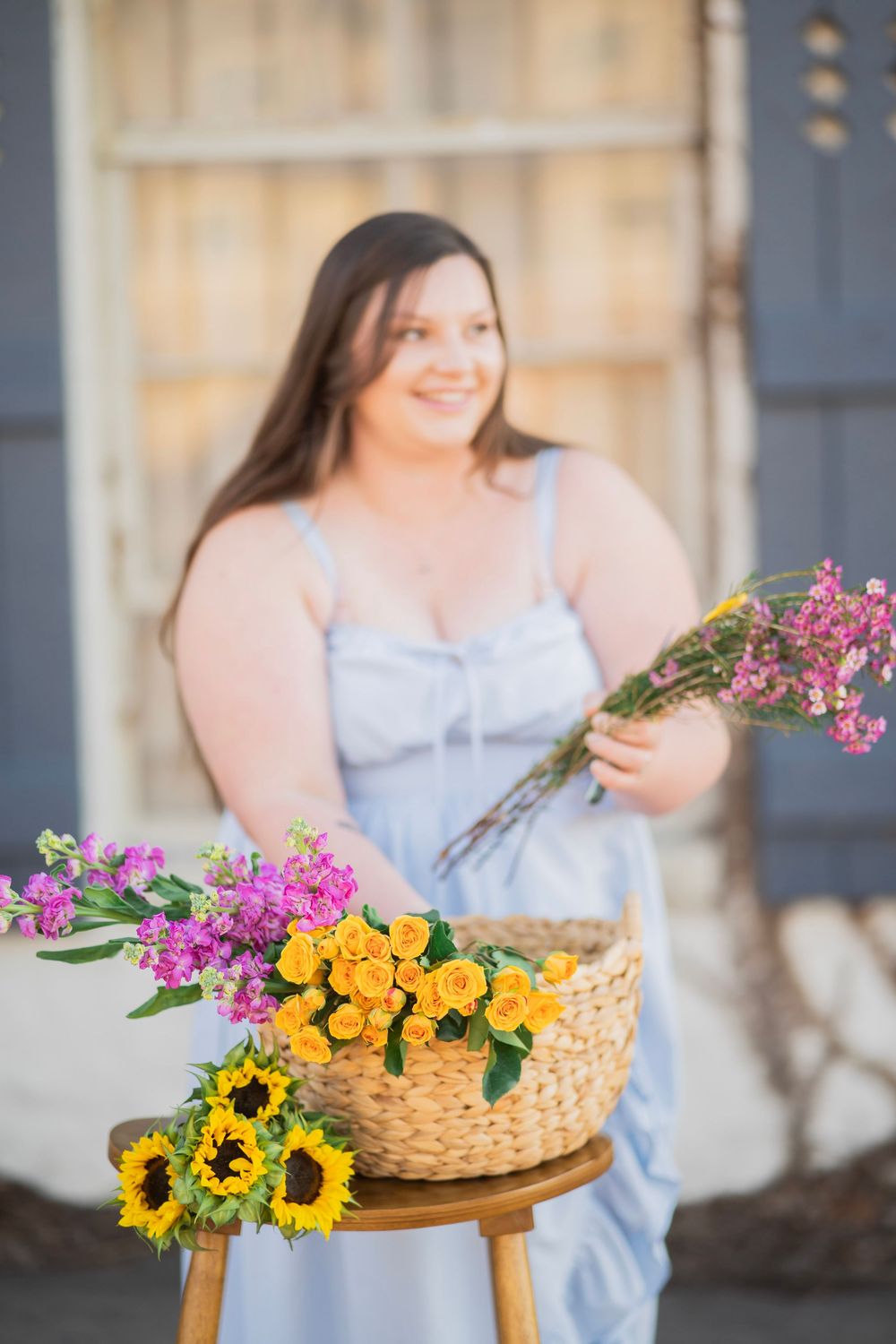 Smiling woman in blue dress holding pink wildflowers behind wicker basket of sunflowers and roses outdoors.