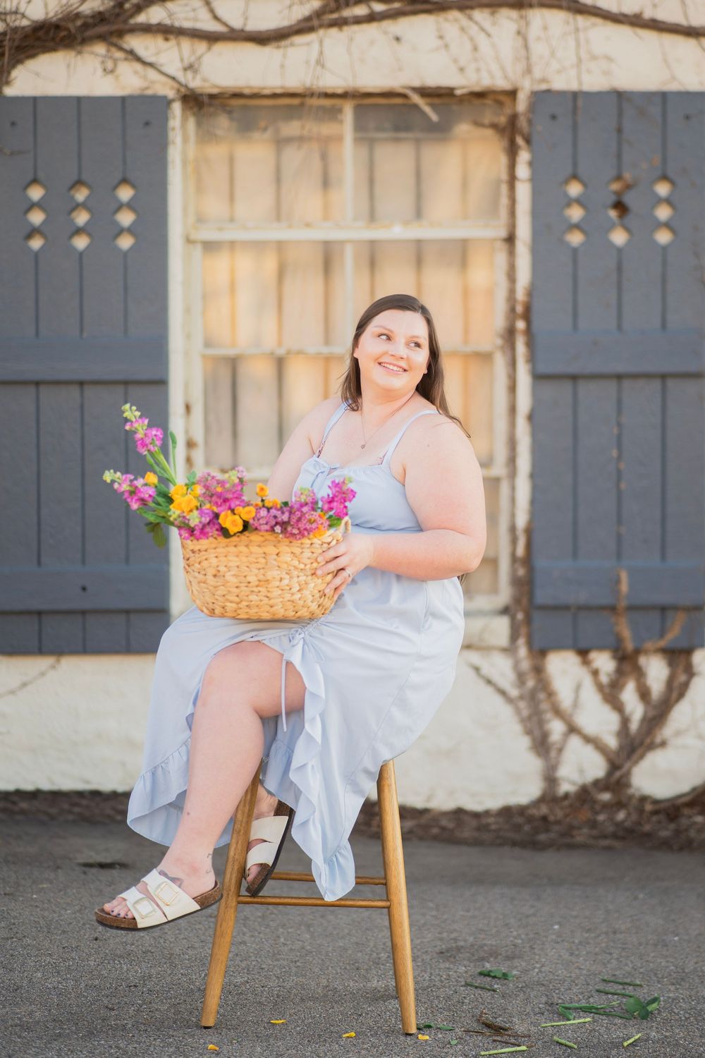Woman in light blue dress sitting on stool holding colorful flower basket outdoors.