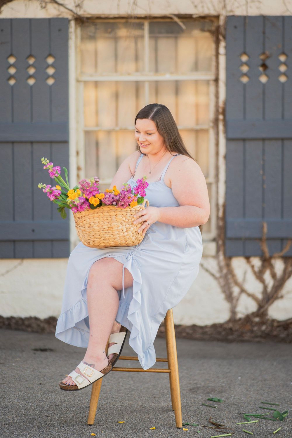Smiling woman in blue dress looking down at vibrant flower basket while seated on stool.
